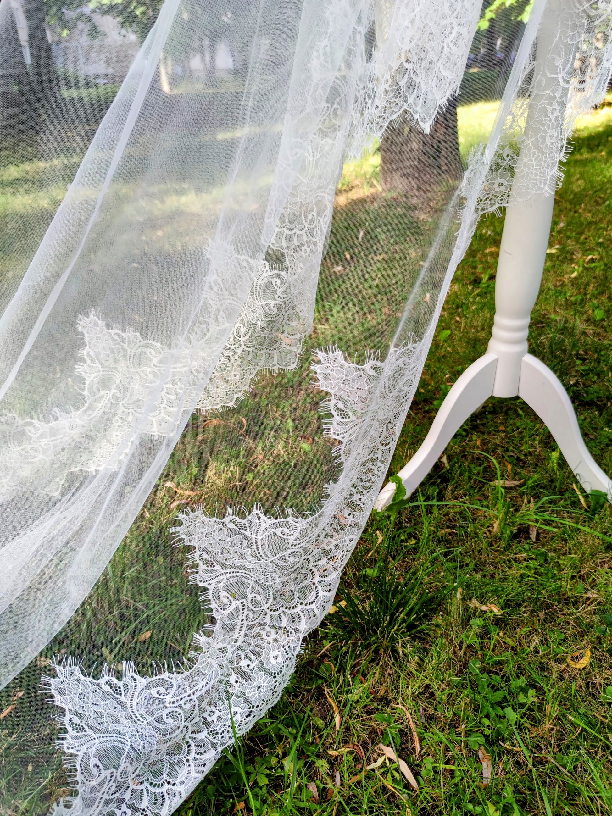 Chantilly lace mantilla veil with blusher draped over a stand, showcasing intricate light ivory lace details in an outdoor setting.