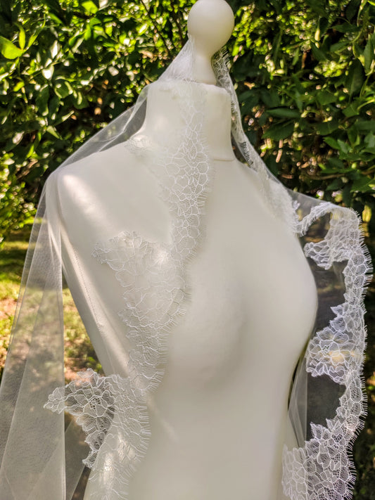 Chantilly lace mantilla veil in white displayed on mannequin against a green outdoor background.