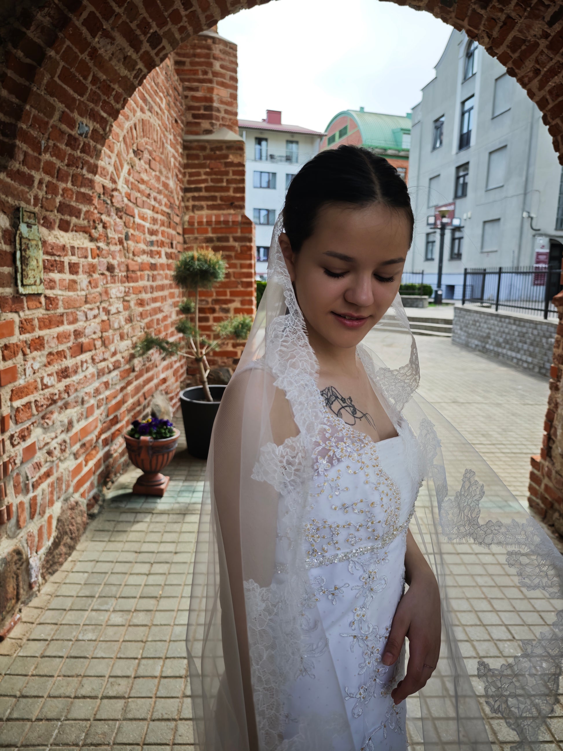Bride under a brick archway wearing the light ivory mantilla veil draped over one shoulder, scalloped floral lace edge
