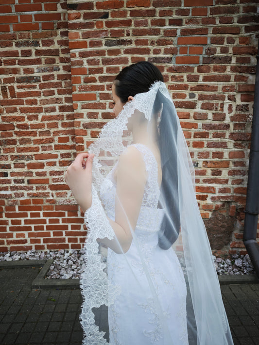 Bride in profile by a brick wall holding the scalloped floral Chantilly lace edge of The Laima single-tier mantilla veil