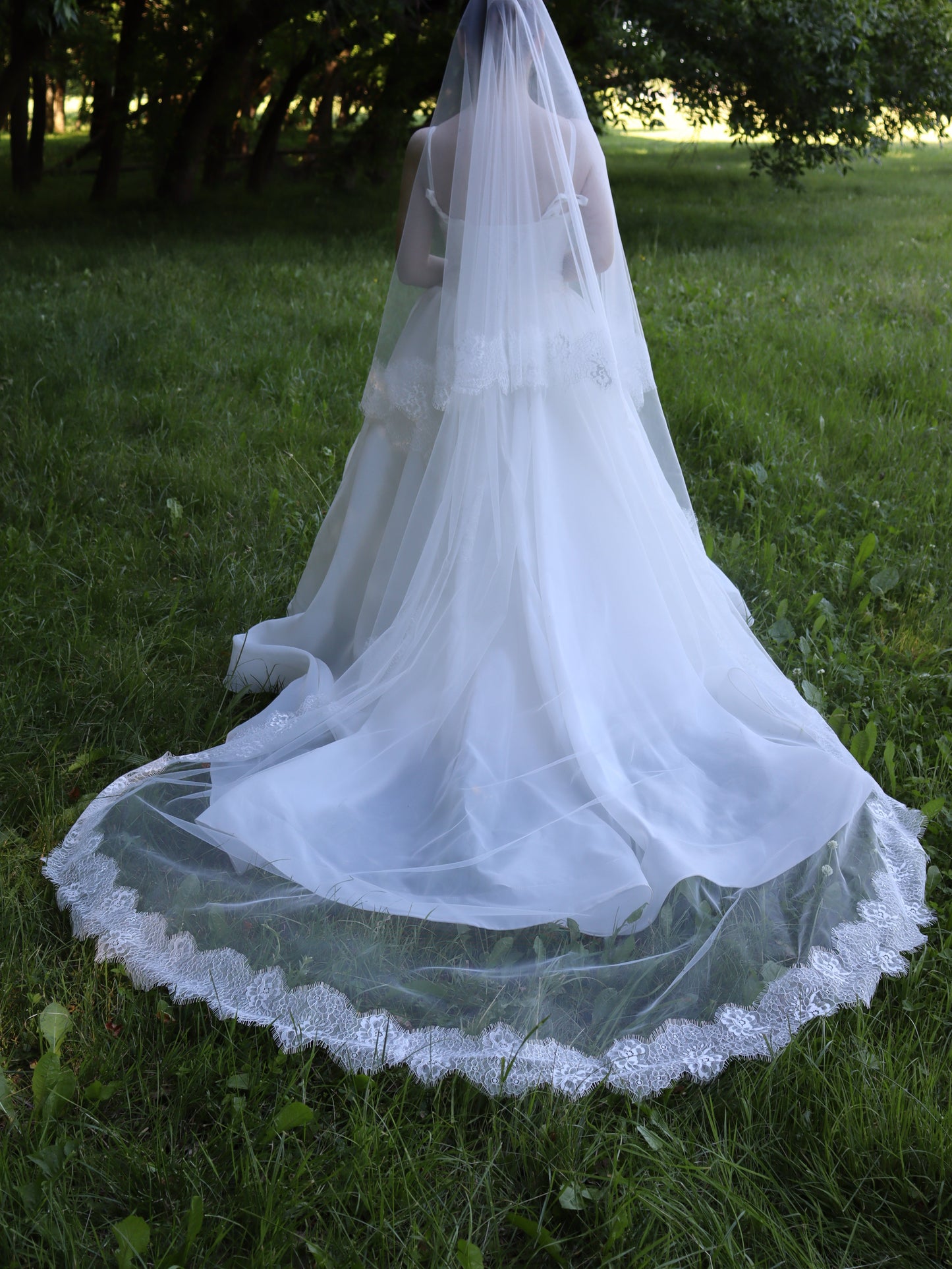 Back view of an ivory two-tier wedding veil with hand-sewn floral lace edge, cathedral length train pooled on the grass