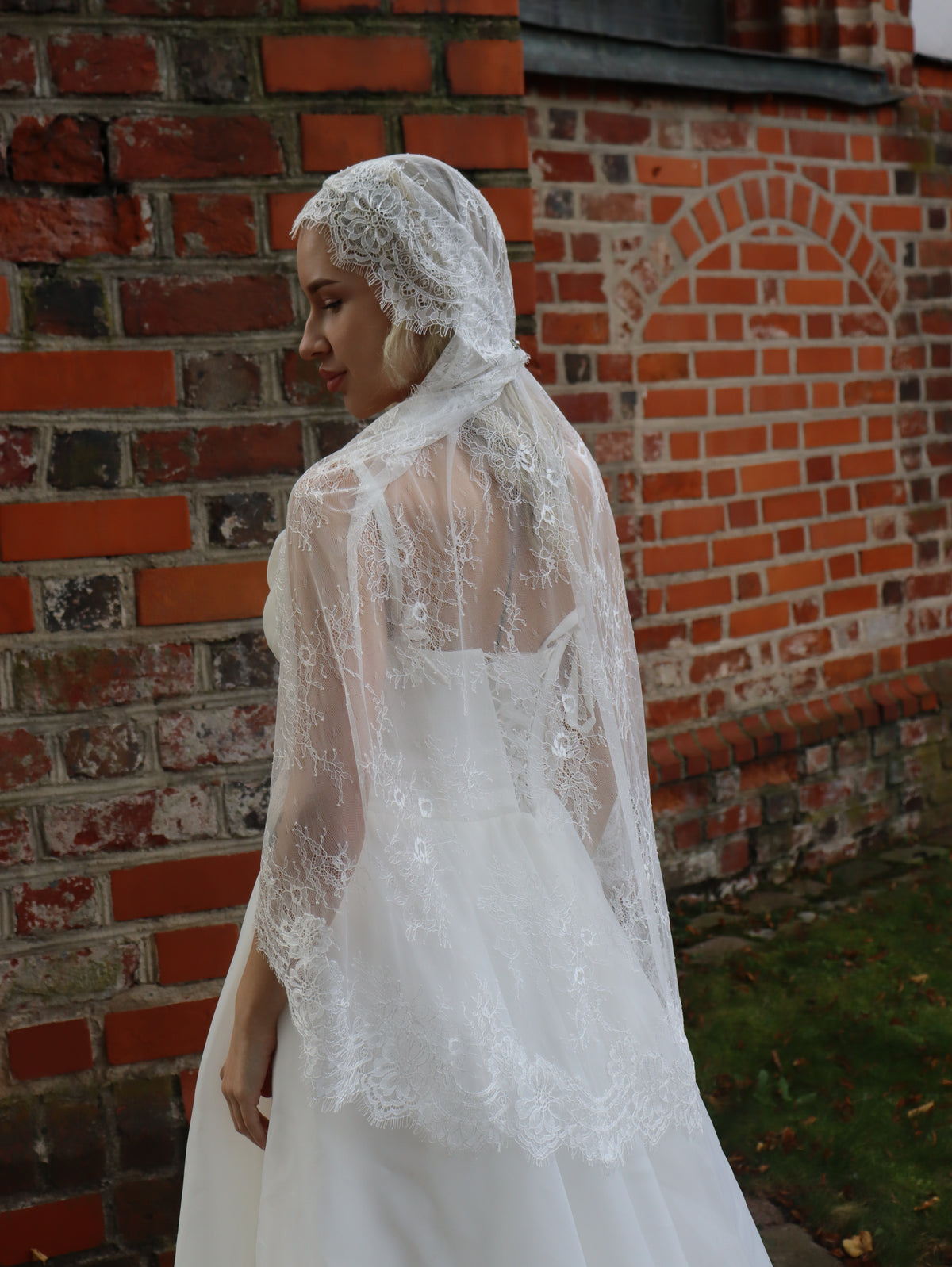 Full lace Juliet cap veil in ivory Chantilly lace, back view, worn by a bride against a red brick wall