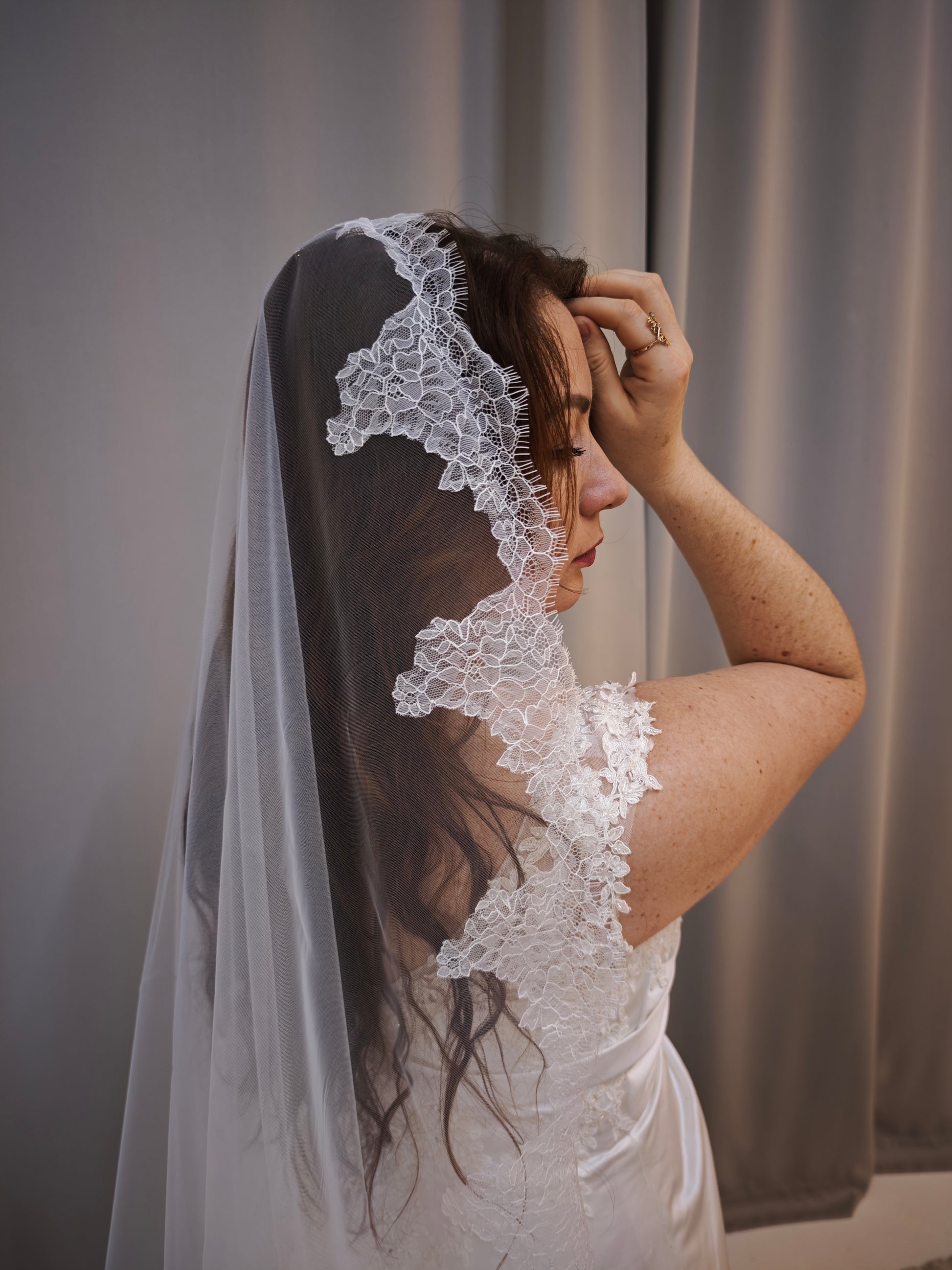 Side profile of a bride indoors with the mantilla veil draped over the head, scalloped floral Chantilly lace framing the face
