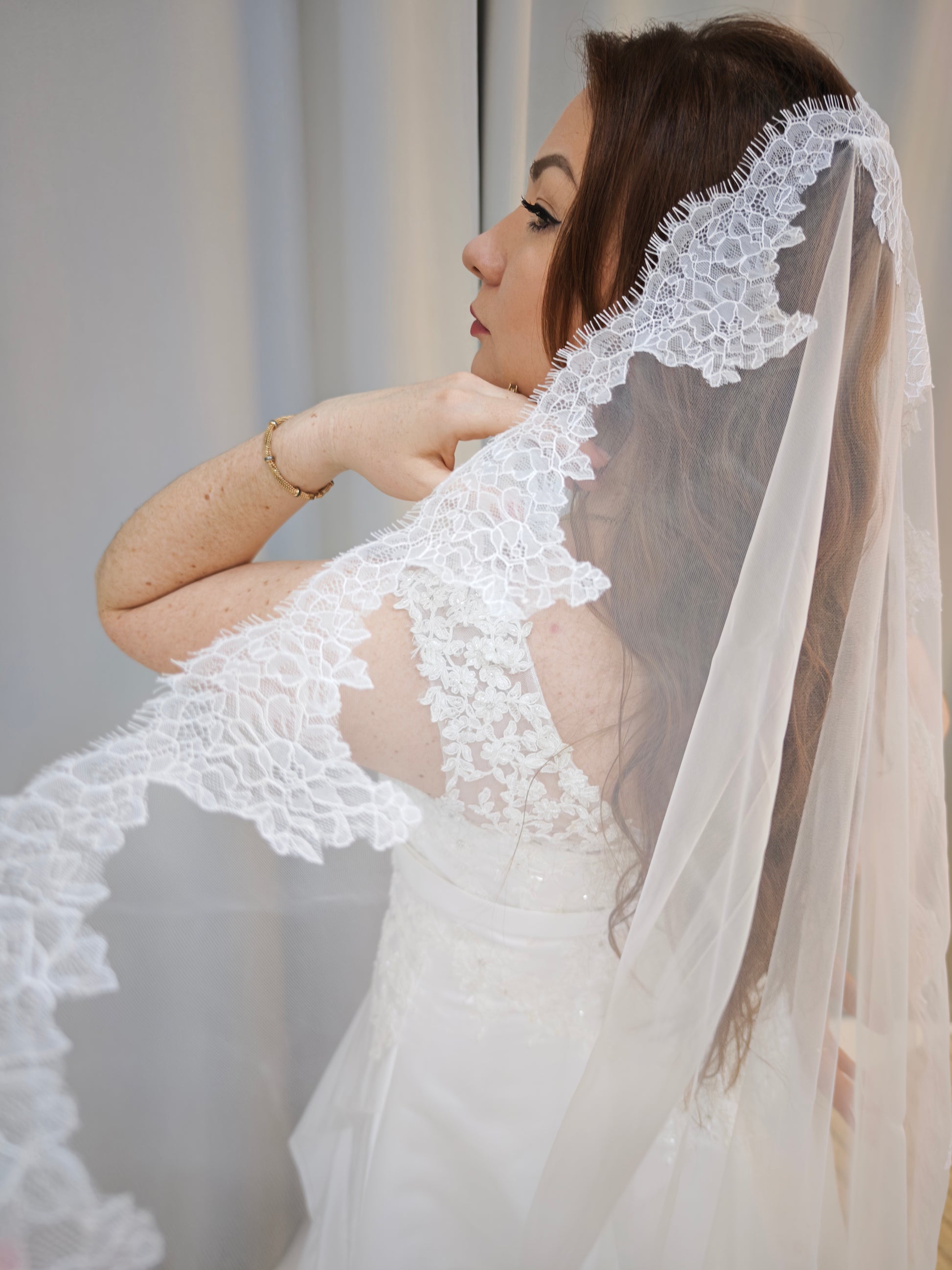 Bride holding the scalloped floral Chantilly lace edge of the light ivory single-tier mantilla veil across the shoulder